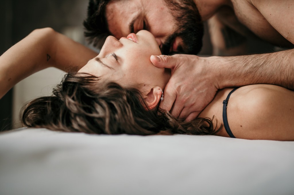 Side photo of a beautiful couple in bed, soft and sensual. Handsome bearded guy on top of beautiful brunette kissing her neck while her eyes are closed. The concept of making love